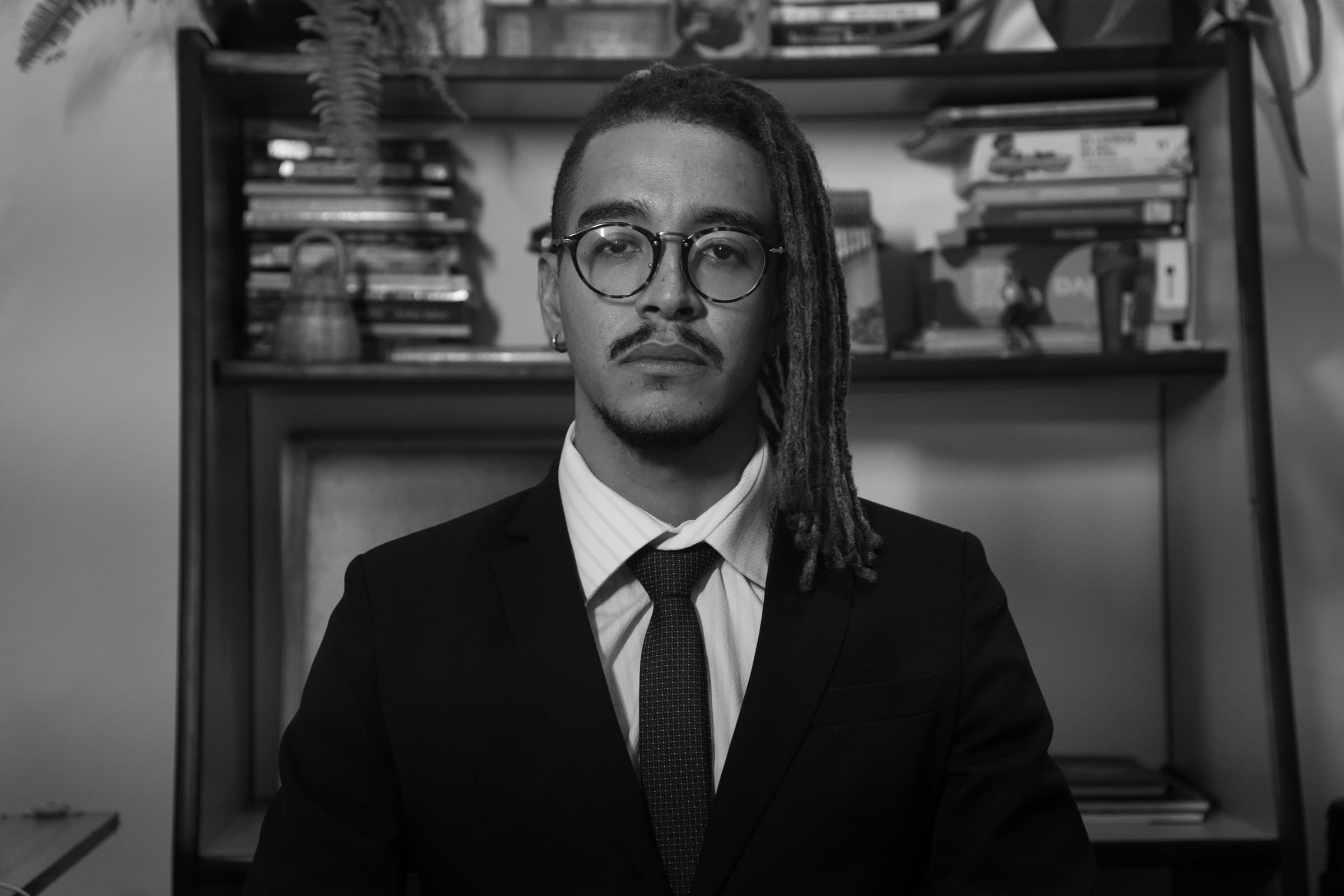 Thiago Farias em preto e branco, sentado diante da estante do escritório